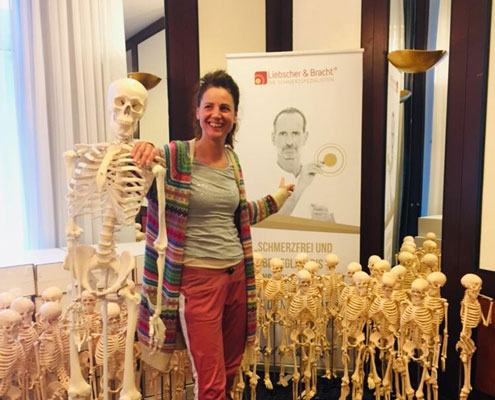 Sibylle Truckenbrodt ausgebildet bei Liebscher & Bracht zum Schmerzspezialist Sibylle Truckenbrodt neben einem Skelett und Aufsteller von Liebscher & Bracht Schmerztherapie